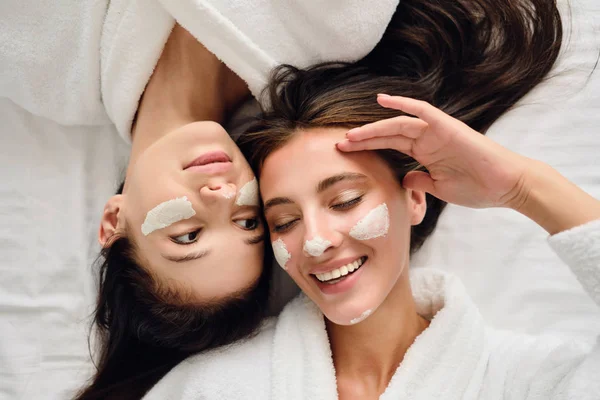 Dos mujeres jóvenes bastante sonrientes con cabello oscuro en batas blancas felizmente tumbadas cerca en la cama con máscara cosmética en las caras en el acogedor hotel moderno —  Fotos de Stock