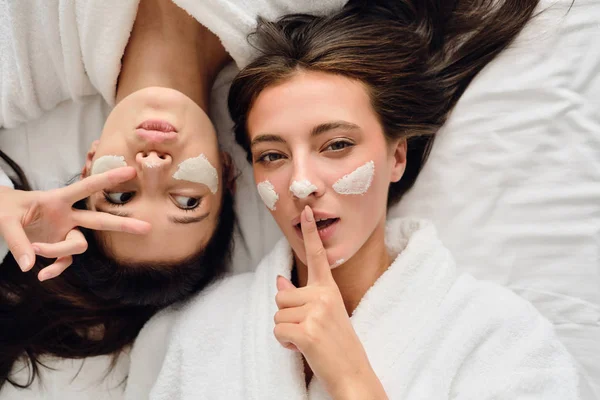 Duas jovens mulheres atraentes com cabelos escuros em roupões de banho brancos deitados perto da cama com máscara cosmética em rostos olhando atenciosamente na câmera juntos no moderno hotel acolhedor — Fotografia de Stock