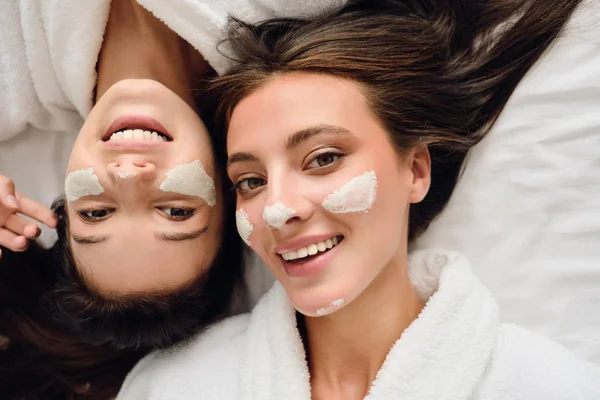 Due giovani donne bellissime con i capelli scuri in accappatoi bianchi sdraiati vicino a letto con maschera cosmetica sui volti felicemente guardando insieme in macchina fotografica in hotel moderno — Foto Stock