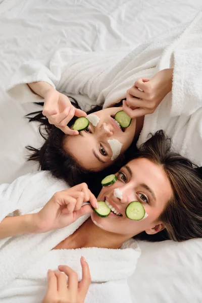 Twee jonge aantrekkelijke vrouwen met donker haar in witte badjassen met cosmetische masker en plakjes komkommer op gezichten liggend in bed dromerige op zoek in de camera samen in het moderne hotel — Stockfoto