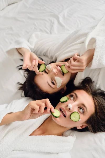 Twee jonge mooie vrouwen met donker haar in witte badjassen liggend in bed met cosmetische masker op gezichten dromerige het eten van plakjes komkommer samen in modern gezellig hotel — Stockfoto