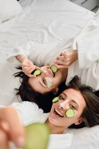 Duas jovens mulheres bonitas com cabelos escuros em roupões de banho brancos com máscara cosmética e fatias de pepino em rostos deitados na cama alegremente olhando para a câmera juntos no hotel moderno — Fotografia de Stock