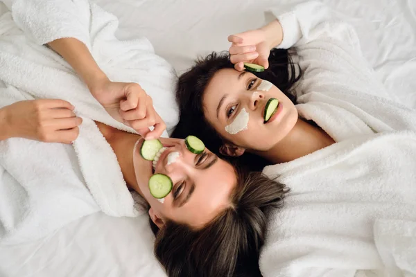 Dos mujeres hermosas jóvenes con cabello oscuro en batas blancas con máscara cosmética y rodajas de pepino en las caras que yacen en la cama mirando en la cámara juntos en el hotel moderno —  Fotos de Stock