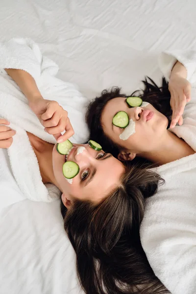 Twee jonge mooie vrouwen met donker haar in witte badjassen met cosmetische masker en plakjes komkommer op gezichten liggend in bed spelvol op zoek in de camera in modern gezellig hotel — Stockfoto