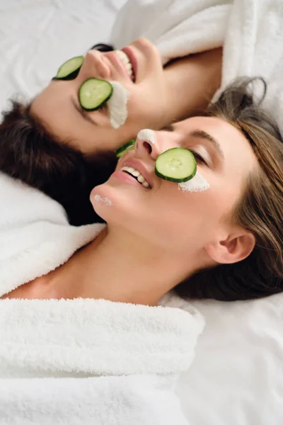 Dos jóvenes y atractivas mujeres sonrientes con cabello oscuro en batas blancas con máscara cosmética y rodajas de pepino en las caras felizmente tumbadas en la cama en el moderno y acogedor hotel —  Fotos de Stock