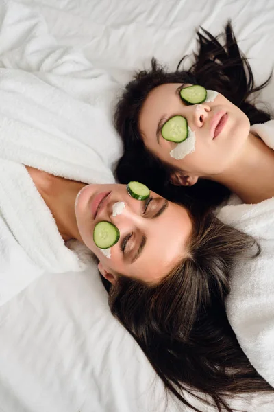 Two young beautiful women with dark hair in white bathrobes with cosmetic mask and slices of cucumber on faces dreamily lying in bed in modern hotel — Stock Photo, Image