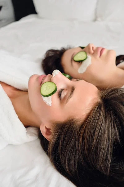 Dua wanita cantik dengan rambut gelap dengan jubah mandi putih dengan topeng kosmetik dan irisan mentimun di wajah berbaring di tempat tidur di hotel modern — Stok Foto