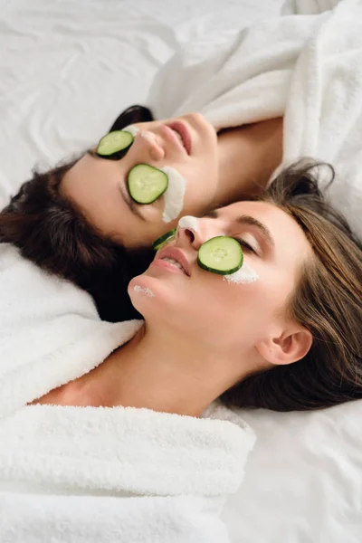 Dos mujeres sensuales atractivas jóvenes con cabello oscuro en batas blancas con máscara cosmética y rodajas de pepino en las caras acostadas en la cama en el hotel moderno —  Fotos de Stock