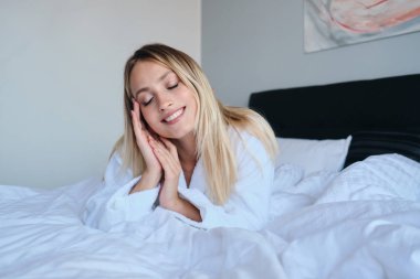 Beyaz bornoz genç çekici gülümseyen sarışın kadın rüya gibi modern rahat otelde yumuşak yatakta yatan gözleri kapanış