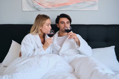 Genç esmer adam ve beyaz bornoz güzel sarışın kadın hayalrüya birlikte kırmızı şarap içme. Güzel çift modern rahat otelde gözlük ile yatakta yalan