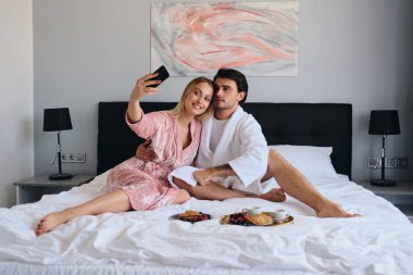 Beyaz bornoz genç esmer adam ve pembe kadife cüppe güzel sarışın kadın mutlu cep telefonu fotoğraf çekmek. Modern rahat otelde yatakta lezzetli kahvaltı yapıyor çekici çift