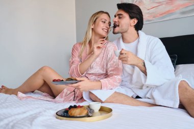 Beyaz bornoz genç esmer adam elinde kahve fincan tutan pembe kadife bornoz güzel sarışın kadın onu besleyen süre. Modern rahat otelde yatakta lezzetli kahvaltı yapıyor çekici çift