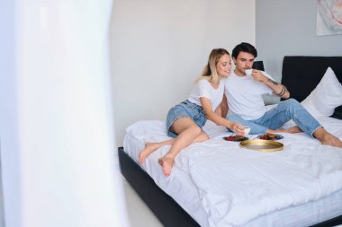 Yakışıklı esmer adam ve güzel sarışın kadın hayalrüya birlikte kahve içme. Modern otelde yatakta lezzetli kahvaltı yapıyor beyaz t-shirt genç çekici çift