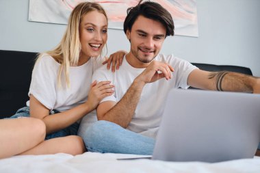 Pretty duygusal sarışın kadın ve çekici gülümseyen esmer adam mutlu birlikte dizüstü bilgisayarda film izliyor. Rahat evde yatakta oturan beyaz t-shirt genç güzel çift