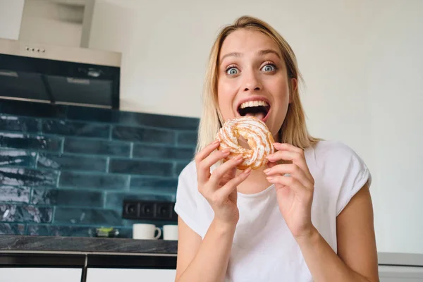 Giovane donna bionda allegra in bianco T-shirt che tiene dolce panino per la prima colazione in mani sorprendentemente guardando in macchina fotografica a casa moderna — Foto Stock