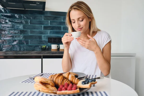 Beyaz t-shirt genç güzel sarışın kadın rüya gibi modern evde kahvaltı ile masada elinde fincan tutan taze kahve kokuyor