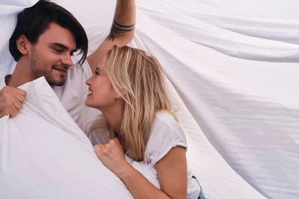 Young smiling brunette man and beautiful blond woman happily looking at each other with pillow in hands lying together under blanket in bed at cozy home — Stock Photo, Image