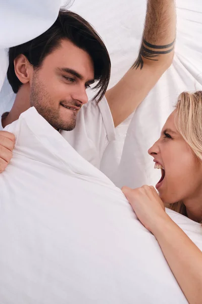 Young brunette man and attractive emotional blond woman looking at each other and fighting for pillow lying together under blanket in bed at home — Stock Photo, Image