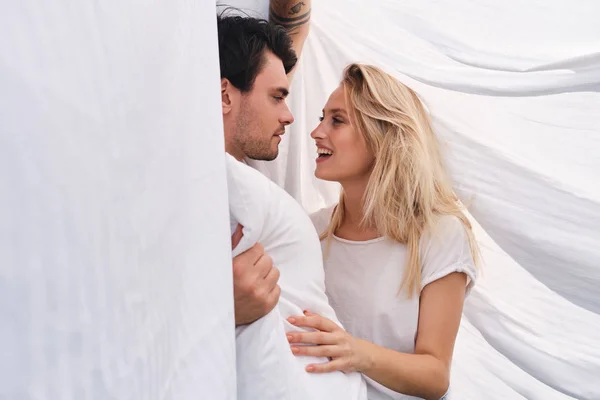 Junge brünette Mann mit Kissen in der Hand verträumt Blick auf schöne lächelnde blonde Frau zusammen unter Decke im Bett zu Hause liegen — Stockfoto