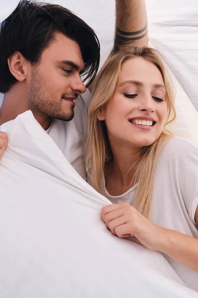 Young attractive brunette man dreamily looking at pretty smiling blond woman lying together under blanket in bed at cozy home — Stock Photo, Image