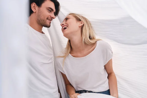 Jonge lachende brunette man en mooie blonde vrouw in wit T-shirts gelukkig kijken naar elkaar zittend onder deken in bed bij Cozy Home — Stockfoto