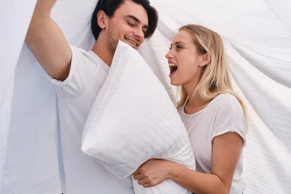 Schöne brünette Mann und ziemlich fröhliche blonde Frau mit weißem Kissen in den Händen glücklich sitzend und mit Decke im Bett zu Hause — Stockfoto