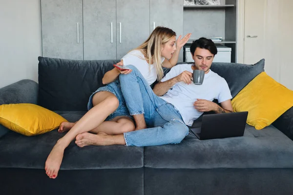 Angry blond woman with cellphone in hand emotionally talking to brunette man with cup of coffee and laptop. Young couple sitting on sofa at modern home