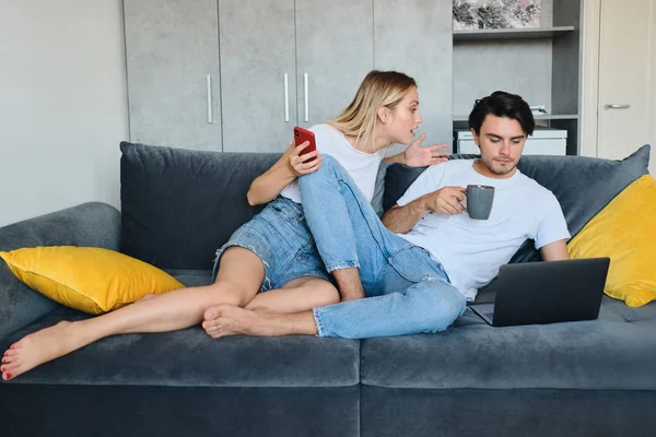 Serious blond woman with cellphone in hand emotionally talking to calm brunette man with cup of coffee and laptop. Young couple in bad mood sitting on sofa at modern home — Stock Photo, Image