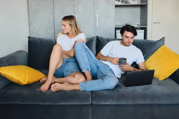 Angry blond woman sadly looking aside while brunette man with cup of coffee thoughtfully using laptop. Young couple sitting on sofa at modern home — Stock Photo, Image
