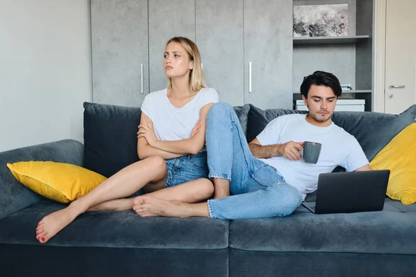 Young offended blond woman sadly looking aside while brunette man with cup of coffee in hand thoughtfully working on laptop. Couple in bad mood sitting on sofa in modern home — Stock Photo, Image