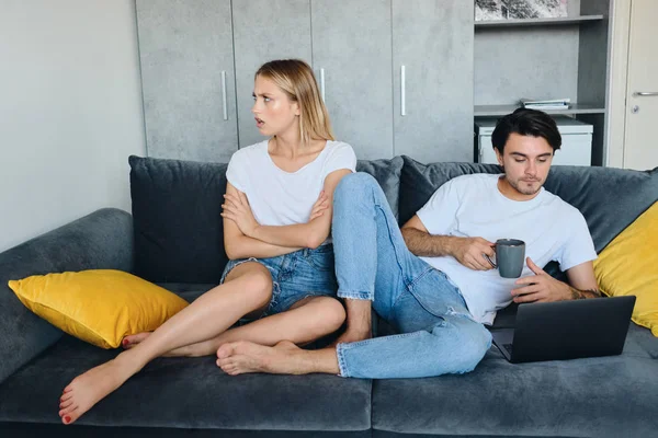 Offended blond woman sadly looking aside while brunette man with cup of coffee in hand thoughtfully using laptop. Young couple in bad mood sitting on sofa in modern home — Stock Photo, Image