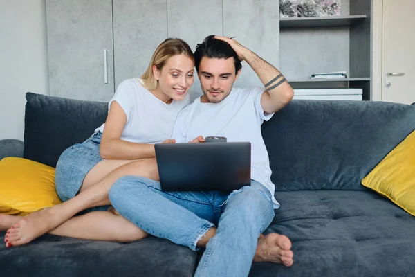 Pretty smiling blond woman and attractive brunette man happily using laptop together. Young beautiful couple sitting on sofa at modern cozy home — Stock Photo, Image