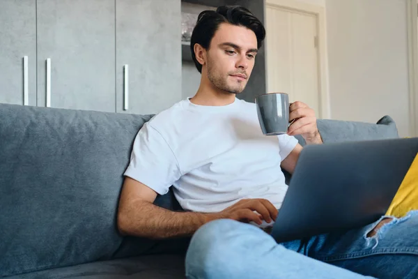 Joven morena atractiva hombre en camiseta blanca sosteniendo la taza en la mano cuidadosamente trabajando en el ordenador portátil sentado en el sofá en el hogar moderno — Foto de Stock