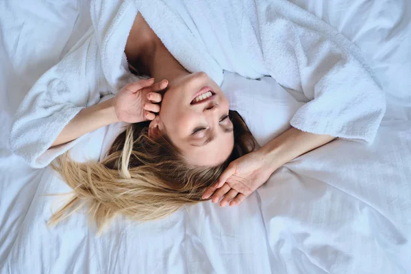 Joven atractiva mujer rubia sonriente en albornoz blanco felizmente cerrando los ojos tumbado en una cama suave en el moderno y acogedor hotel —  Fotos de Stock