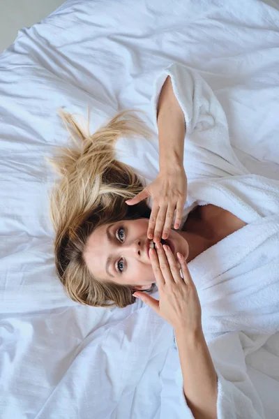 Ung vacker blond kvinna i vit morgonrock som täcker munnen med händer häpligt tittar i kameran liggande i mjuk säng i modernt mysigt hotell — Stockfoto
