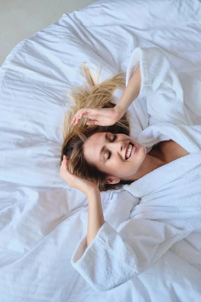 Young beautiful smiling blond woman in white bathrobe dreamily closing eyes lying in soft bed in modern cozy hotel — Stock Photo, Image