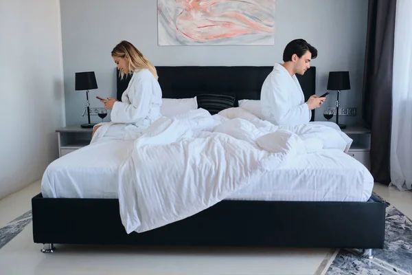 Jeune homme brune réfléchi et jolie femme blonde en peignoirs blancs à l'aide de téléphones cellulaires assis sur les différents côtés du lit dans un hôtel confortable moderne — Photo
