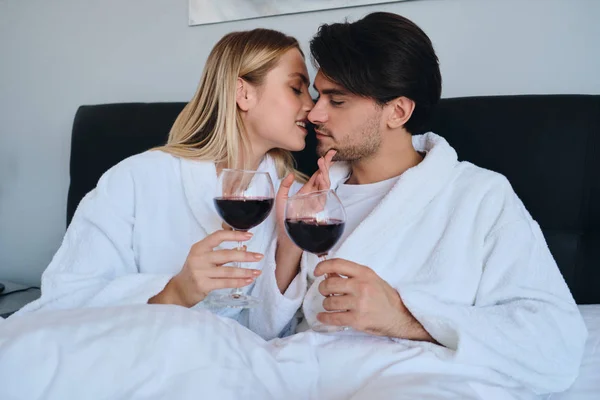 Junger brünetter Mann und hübsche blonde Frau in weißen Bademänteln, die sich verträumt küssen, mit einem Glas Rotwein in der Hand. Schönes Paar liegt im Bett in modernem, gemütlichem Hotel — Stockfoto