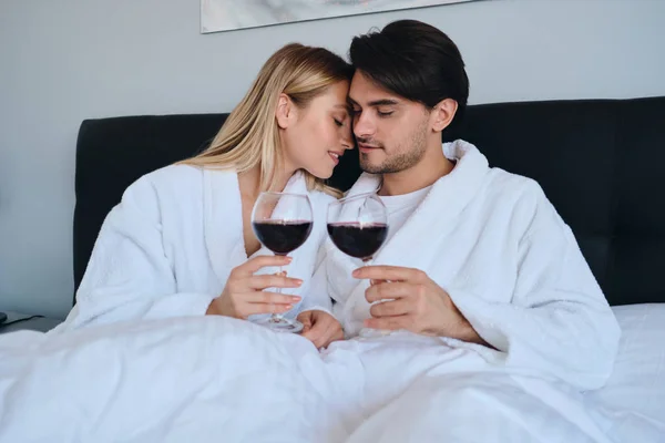 Νέος μελαχρινή άντρας και όμορφη ξανθιά γυναίκα με λευκά μπουρνούζια ονειρικά πίνοντας κόκκινο κρασί μαζί. Όμορφο ζευγάρι που βρίσκεται στο κρεβάτι στο μοντέρνο φιλόξενο ξενοδοχείο — Φωτογραφία Αρχείου