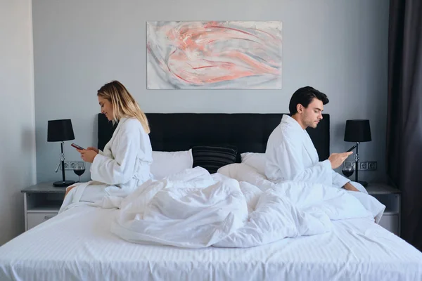Jeune homme brune et jolie femme blonde en peignoirs blancs à l'aide de téléphones cellulaires assis sur différents côtés du lit dans un hôtel confortable moderne — Photo