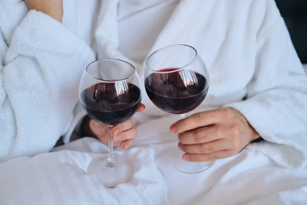 Acercamiento hombre y mujer en batas blancas sosteniendo copas de vino tinto en las manos tumbados en la cama en el moderno y acogedor hotel — Foto de Stock