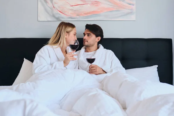 Handsome brunette man and beautiful blond woman in white bathrobes happily talking with glasses of red wine in hands. Young attractive couple lying in bed in modern cozy hotel — Stock Photo, Image