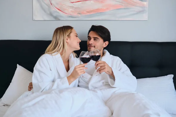 Привабливий брюнетка чоловік і красива блондинка в білих халатах щасливо дивляться один на одного з келихами червоного вина в руках. Молода красива пара лежить в ліжку в сучасному затишному готелі — стокове фото