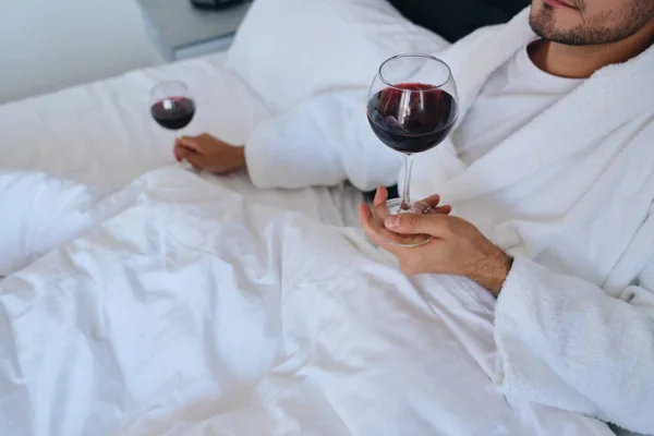 Foto de cerca de un joven atractivo en albornoz blanco acostado en la cama con copas de vino tinto en las manos en el moderno y acogedor hotel — Foto de Stock