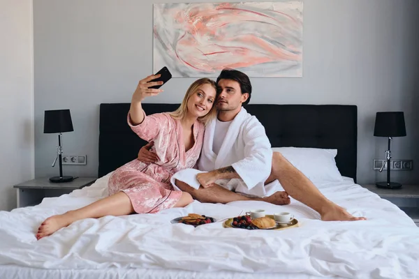 Beyaz bornoz genç esmer adam ve pembe kadife cüppe güzel sarışın kadın mutlu cep telefonu fotoğraf çekmek. Modern rahat otelde yatakta lezzetli kahvaltı yapıyor çekici çift
