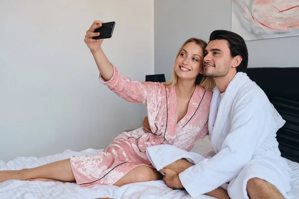 Junge brünette Mann im weißen Bademantel und schöne blonde Frau in rosa Samtgewand glücklich Foto auf dem Handy. Hübsches Paar sitzt zusammen im Bett in modernem, gemütlichem Hotel — Stockfoto