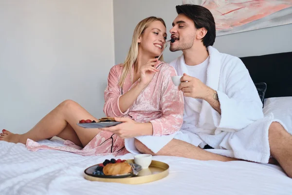 Beyaz bornoz genç esmer adam elinde kahve fincan tutan pembe kadife bornoz güzel sarışın kadın onu besleyen süre. Modern rahat otelde yatakta lezzetli kahvaltı yapıyor çekici çift