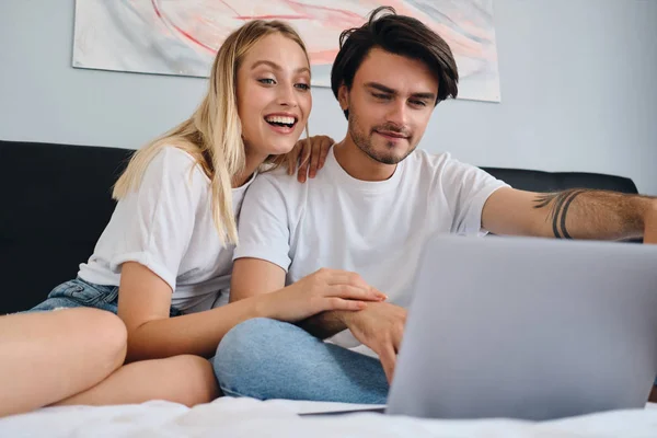 Pretty cheerful blond woman and attractive brunette man happily watching movie on laptop together. Young cute couple in white T-shirts sitting on bed at cozy home — Stock Photo, Image