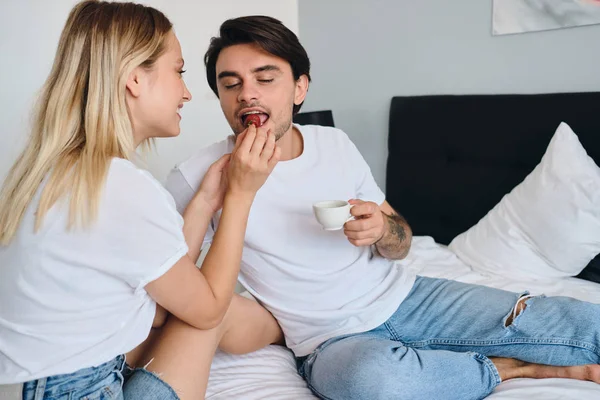 Young brunette man holding cup of coffee in hand dreamily eating strawberry while pretty blond woman feeding him. Beautiful couple in white T-shirts having delicious breakfast in bed in modern hotel
