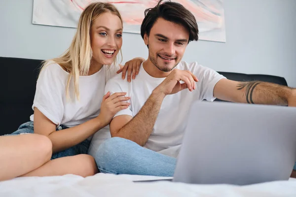 Pretty emotional blond woman and attractive smiling brunette man happily watching movie on laptop together. Young beautiful couple in white T-shirts sitting on bed at cozy home — Stock Photo, Image